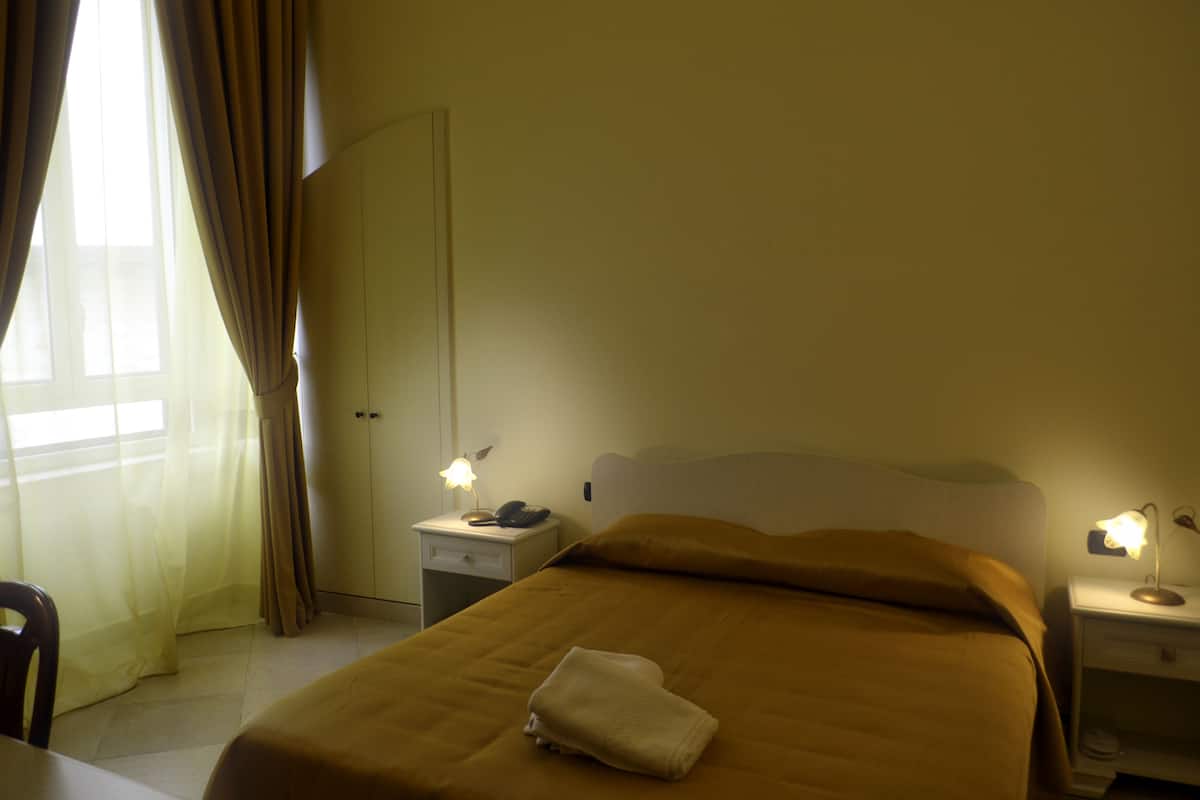 A hotel room with a bed, two bedside tables with lamps, a chair, and a window with curtains.