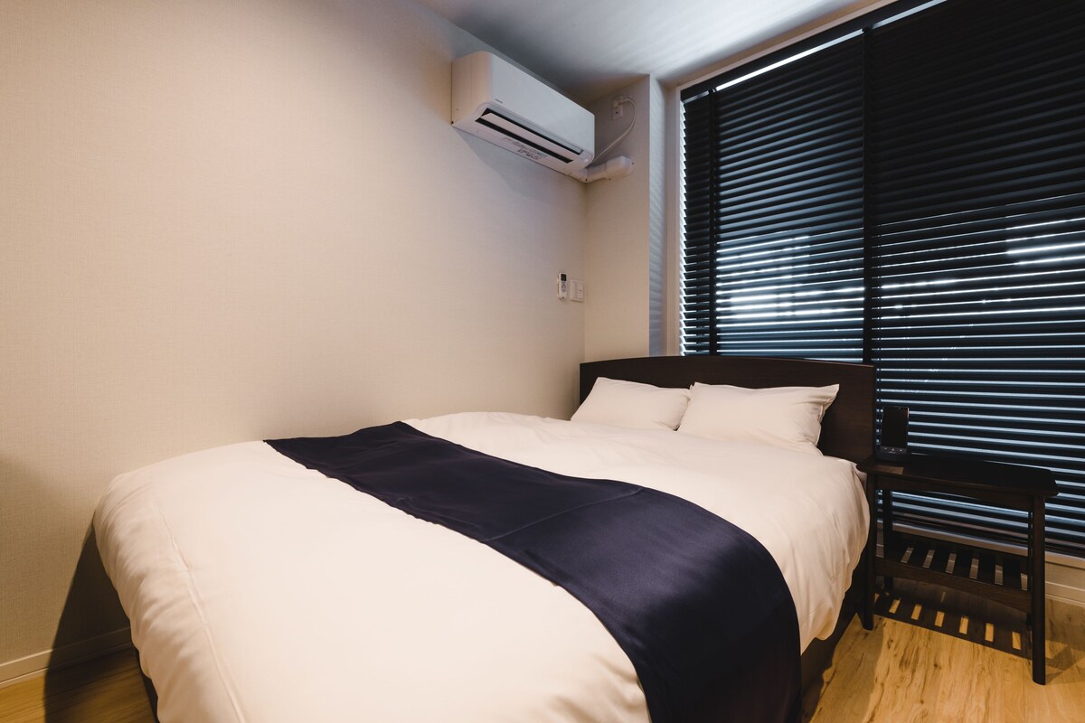 A neatly made bed with a dark headboard, a bedside table, and a window with blinds.