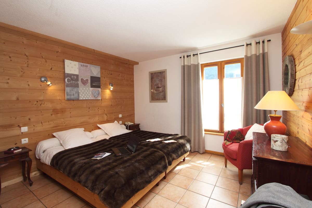 A bedroom with a wooden headboard, a bed with a fur throw, a laptop, a red chair, a lamp, and a window with curtains.
