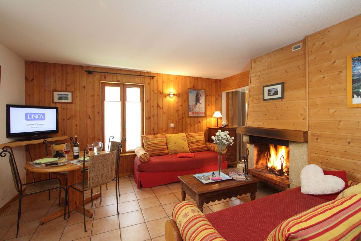 A cozy living room with a fireplace, wooden walls, a sofa, a dining table with chairs, and a television displaying 'DNA'.