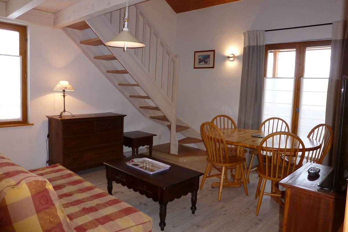 A cozy living room with a wooden staircase, a dining table with chairs, a sofa, and a lamp.