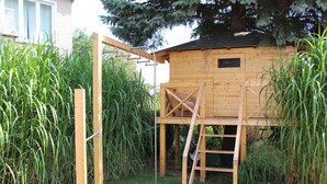 Une cabane en bois, dotée d’une échelle et d’une structure d’escalade, entourée d’herbe haute et d’un arbre.