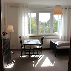 A room with a dining table, chairs, a lamp, a bed, and a window with curtains. - Pension Belger (Blankenfelde-Mahlow)