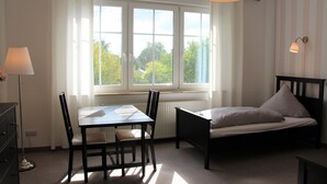 Une pièce comprenant un lit, une table et des chaises. Il y a une fenêtre avec des rideaux et une lampe.