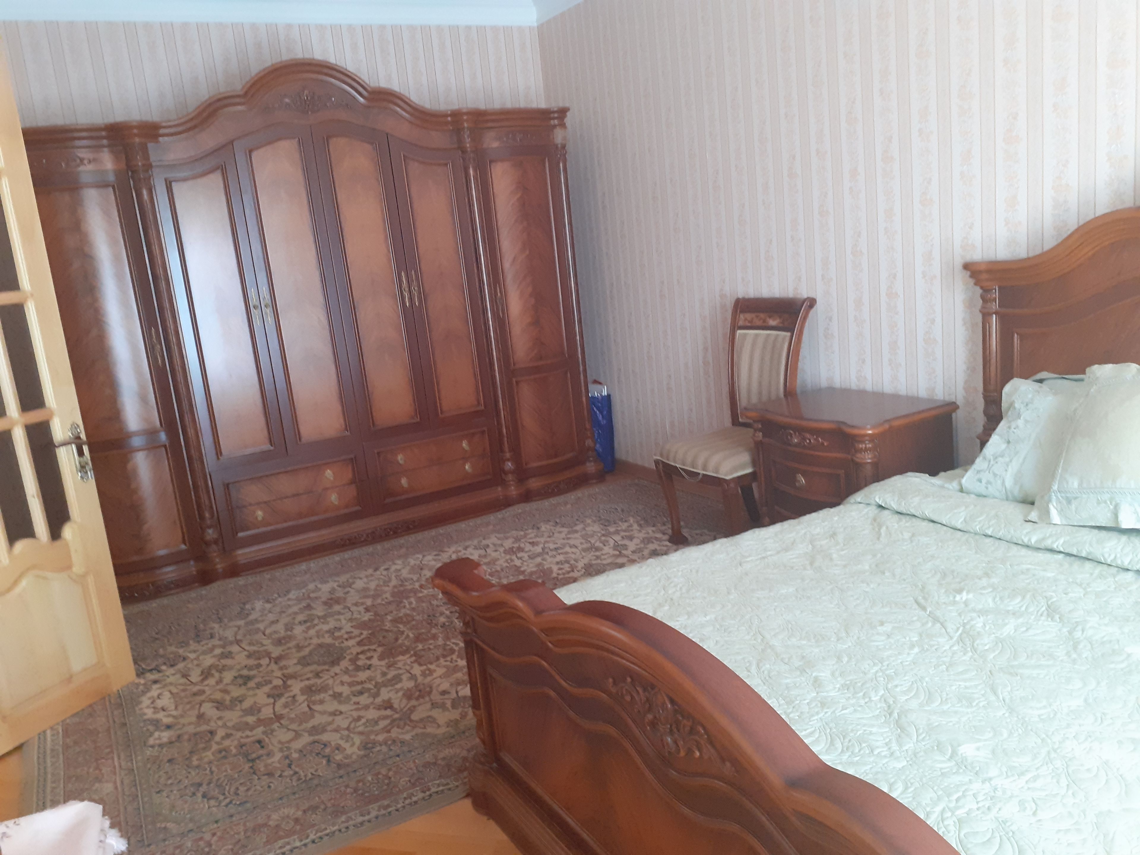 A bedroom with a large wooden wardrobe, a bed with a white quilt, a wooden nightstand, and a chair.