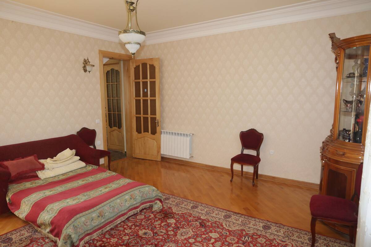 A bedroom with a bed, a red sofa, a wooden chair, a wooden cabinet, and a patterned rug.