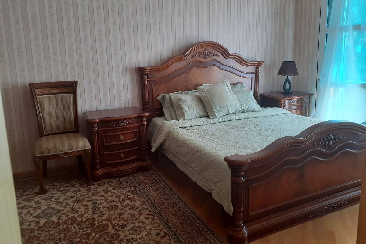A bedroom with a wooden bed, a nightstand, a chair, and a patterned rug.
