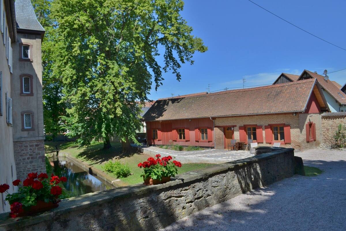 A traditional building with a stone facade, a red-tiled roof, and a small pond with red flowers in the foreground.