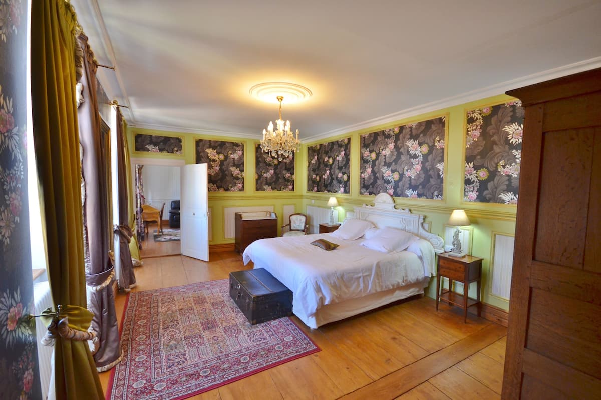 A bedroom with a large bed, a wooden floor, a chandelier, and floral wallpaper.
