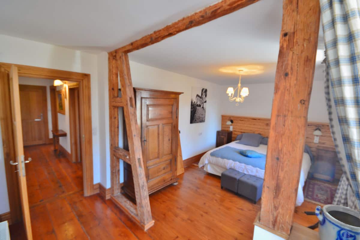 A room with wooden flooring, a bed with a blue blanket, a wooden wardrobe, and a chandelier.