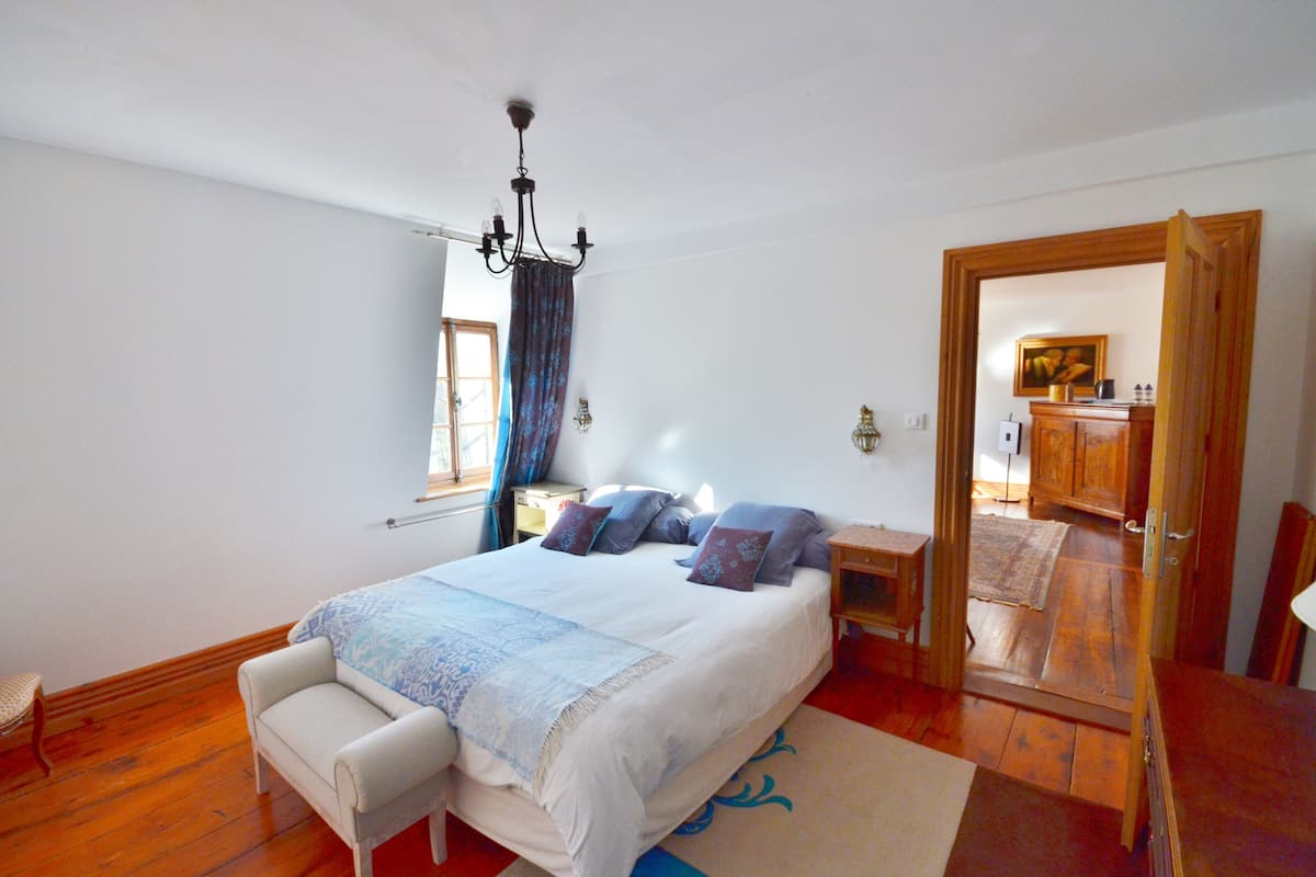 A bedroom with a large bed, a bedside table, a chair, a wooden wardrobe, and a mirror.