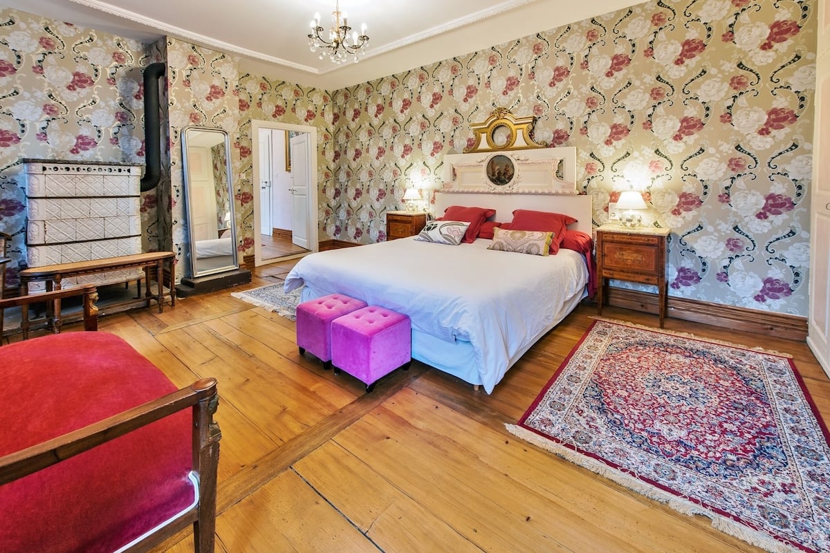 A bedroom with a large bed, a wooden bench, a red chair, a mirror, and a floral wallpaper.