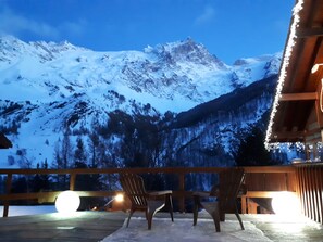 Une terrasse en bois avec deux chaises et une table, d’où l’on peut admirer une chaîne de montagnes enneigées.