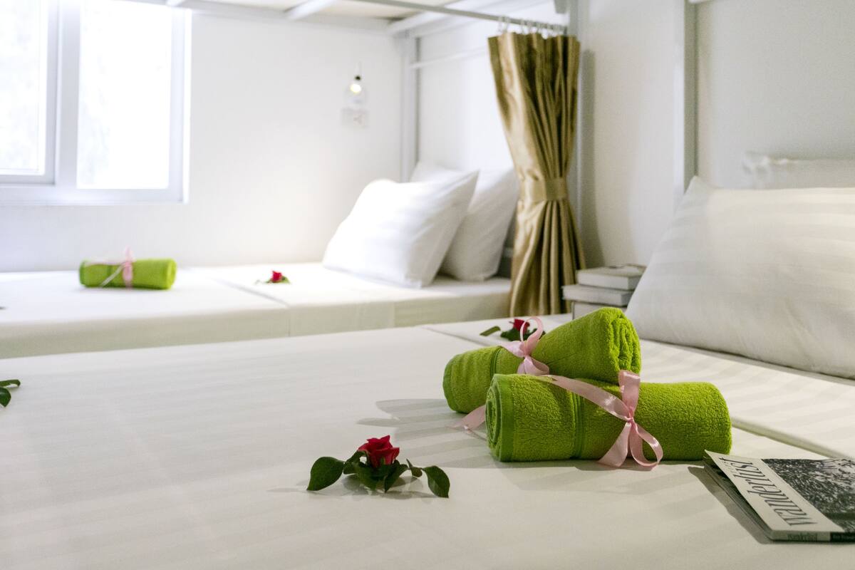 A neatly made bed with green towels, a rose, and a magazine on a white bedspread in a bright room with a window and curtains.