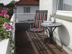 Un balcón con una silla, una mesita, barandilla blanca y una ventana con cortina.