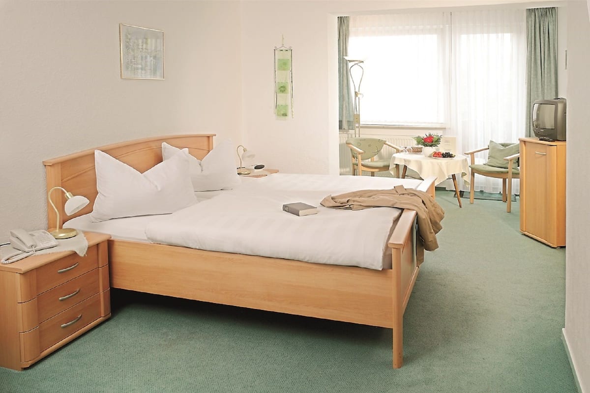Une chambre d’hôtel avec un lit en bois, une table de chevet avec une lampe, un bureau avec une chaise et un téléviseur posé sur un meuble.