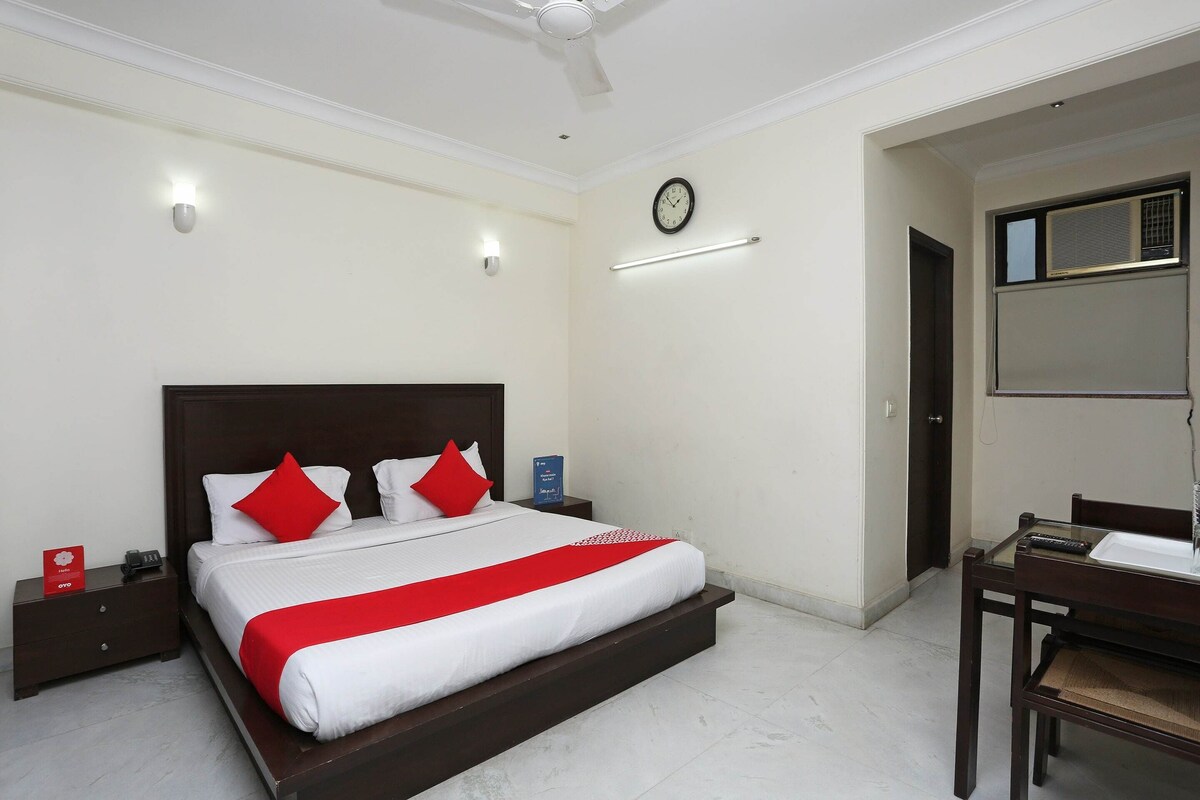 A hotel room with a large bed, a bedside table with a phone and a red sign, a wall clock, and a desk with a chair.