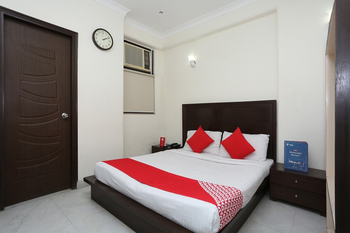 A hotel room with a large bed, a wooden door, a wall clock, a bedside table with a phone, and a sign on the nightstand.