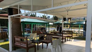 A covered outdoor seating area with multiple tables and chairs, a green umbrella, and a view of a parking lot. - New Iday Hotel (Kusadasi)