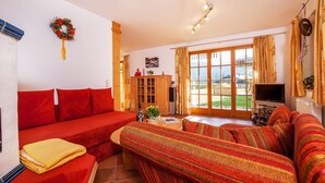 A cozy living room with a red sofa, a striped couch, and a TV. There's a wooden cabinet and a window with curtains. - Evis Wohnung (Reit im Winkl)