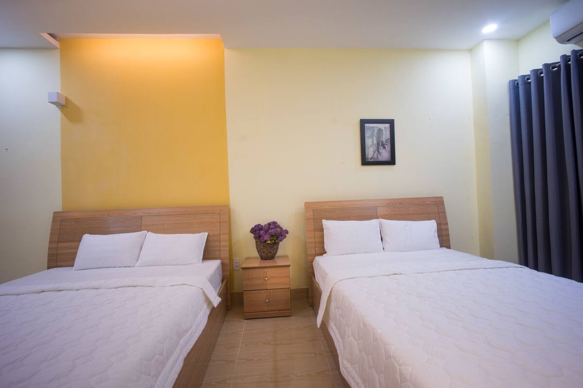 Quarto de hotel com duas camas, uma pequena mesa de cabeceira com um vaso de flores e um quadro na parede.