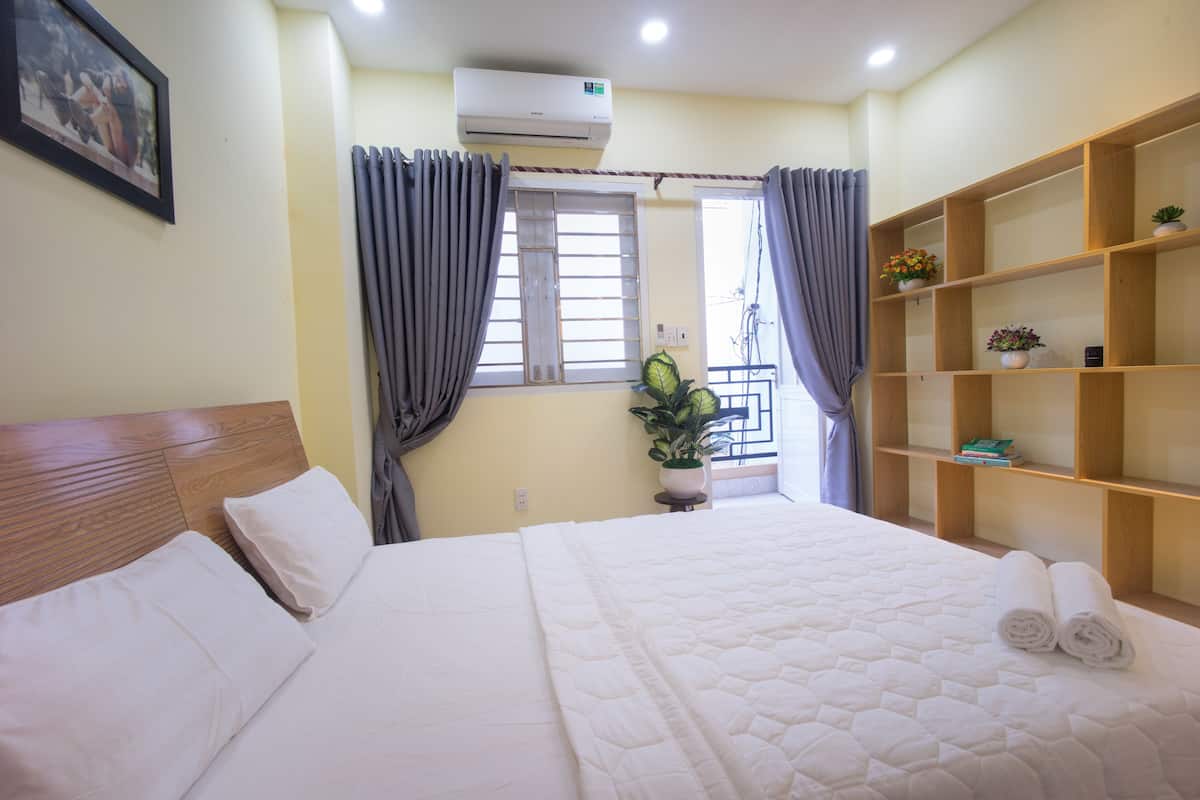 Quarto de hotel com uma cama grande, cabeceira de madeira, janela com cortinas, uma mesinha com uma planta, e uma estante com livros e um vaso de plantas.