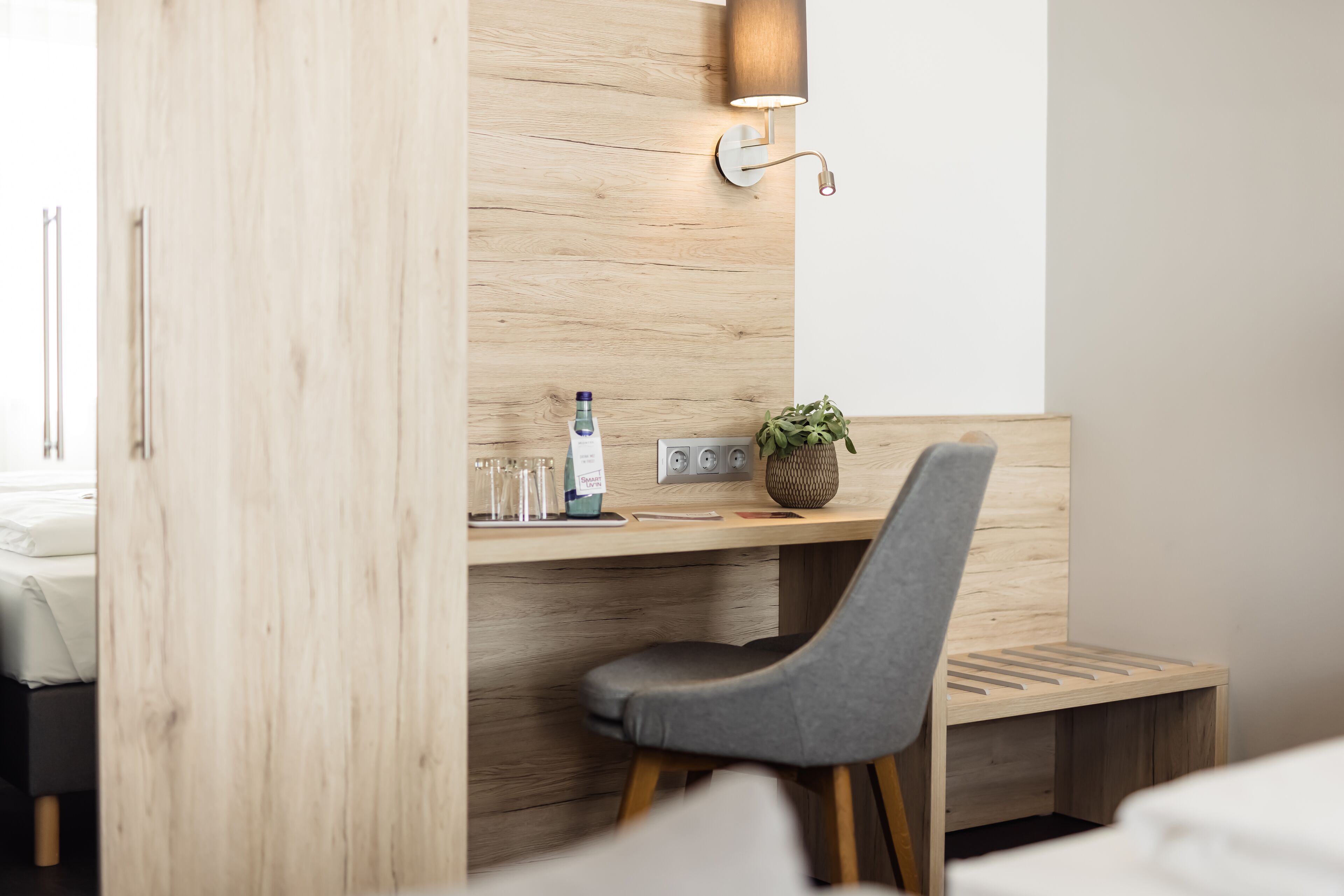A modern hotel room with a wooden desk, a grey chair, a small plant, and a wall-mounted lamp.