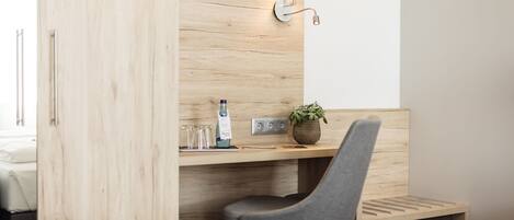 A modern hotel room with a wooden desk, a grey chair, a small plant, and a wall-mounted lamp.