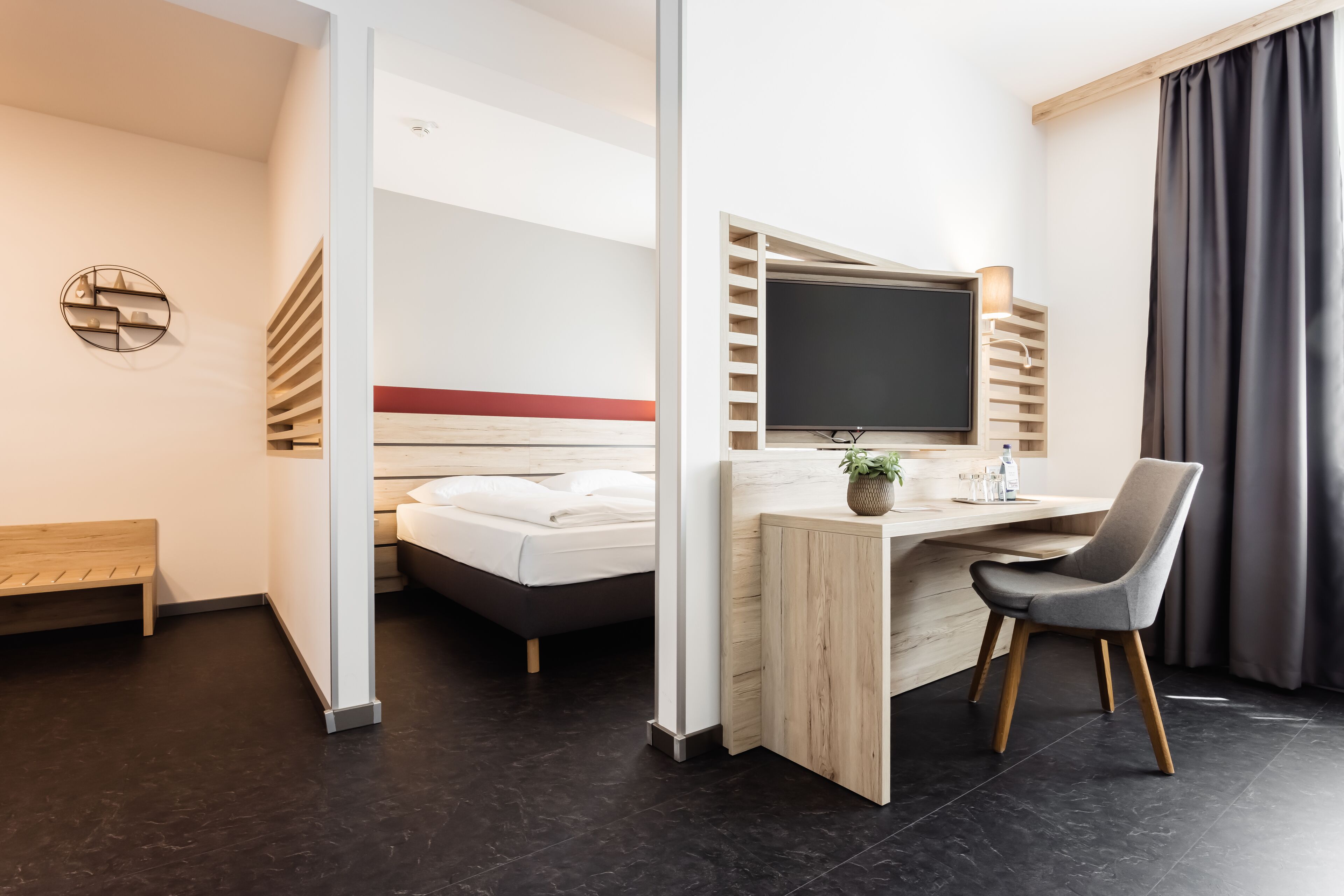 A modern hotel room with a bed, a desk with a chair, a TV, and a window with curtains.