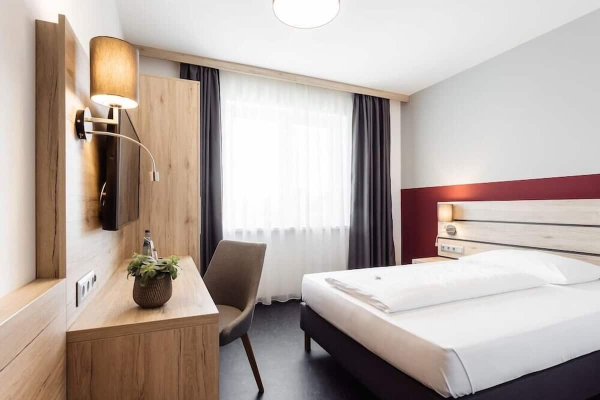 A hotel room with a bed, a desk, a chair, a lamp, and a window with curtains.