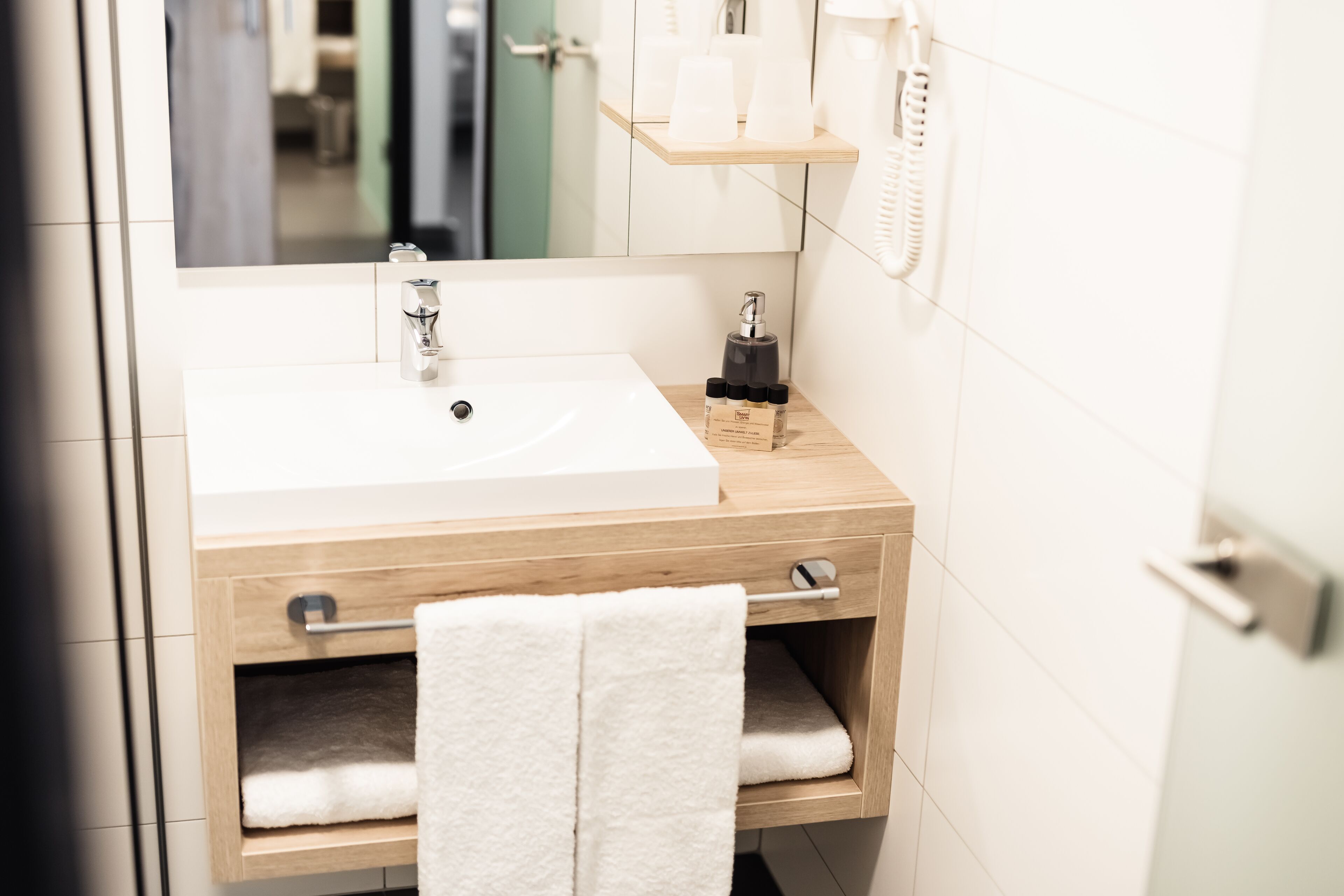 Un bagno con lavandino bianco, mobile contenitore in legno, porta asciugamani e specchio.