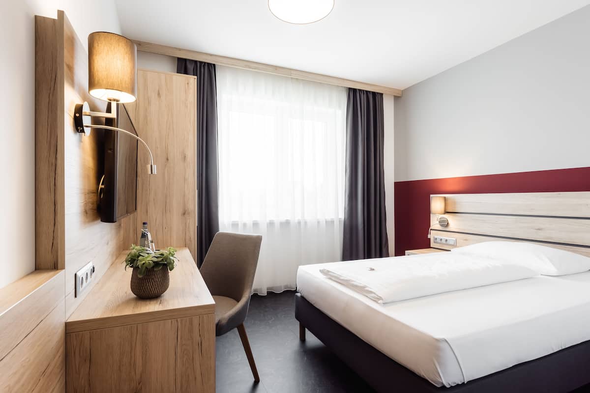 A hotel room with a bed, a desk with a lamp, a chair, and a window with curtains.