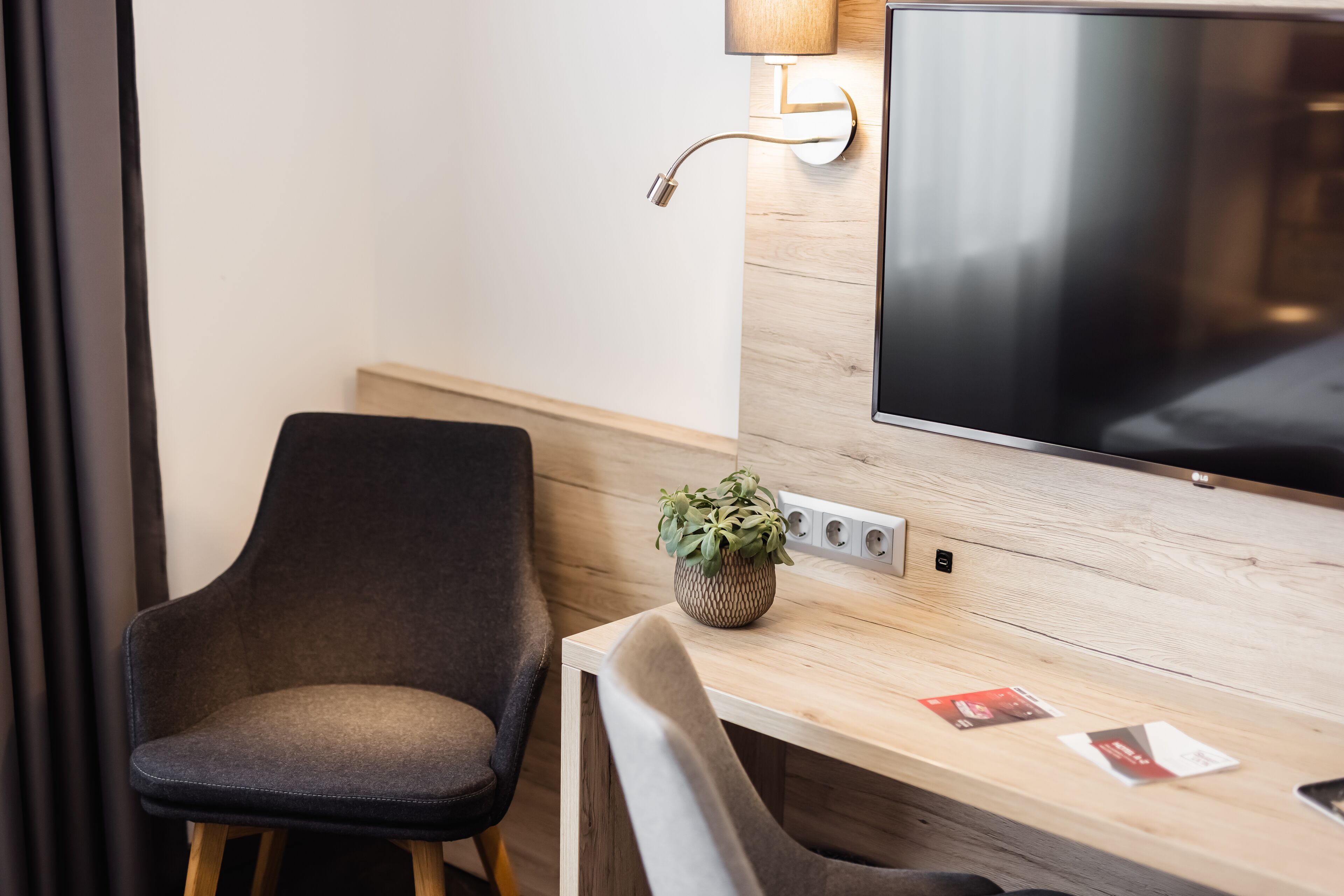 Ein modernes Hotelzimmer mit einem Holztisch, einem flachbildfernseher an der Wand, einer kleinen Topfpflanze und zwei Stühlen.
