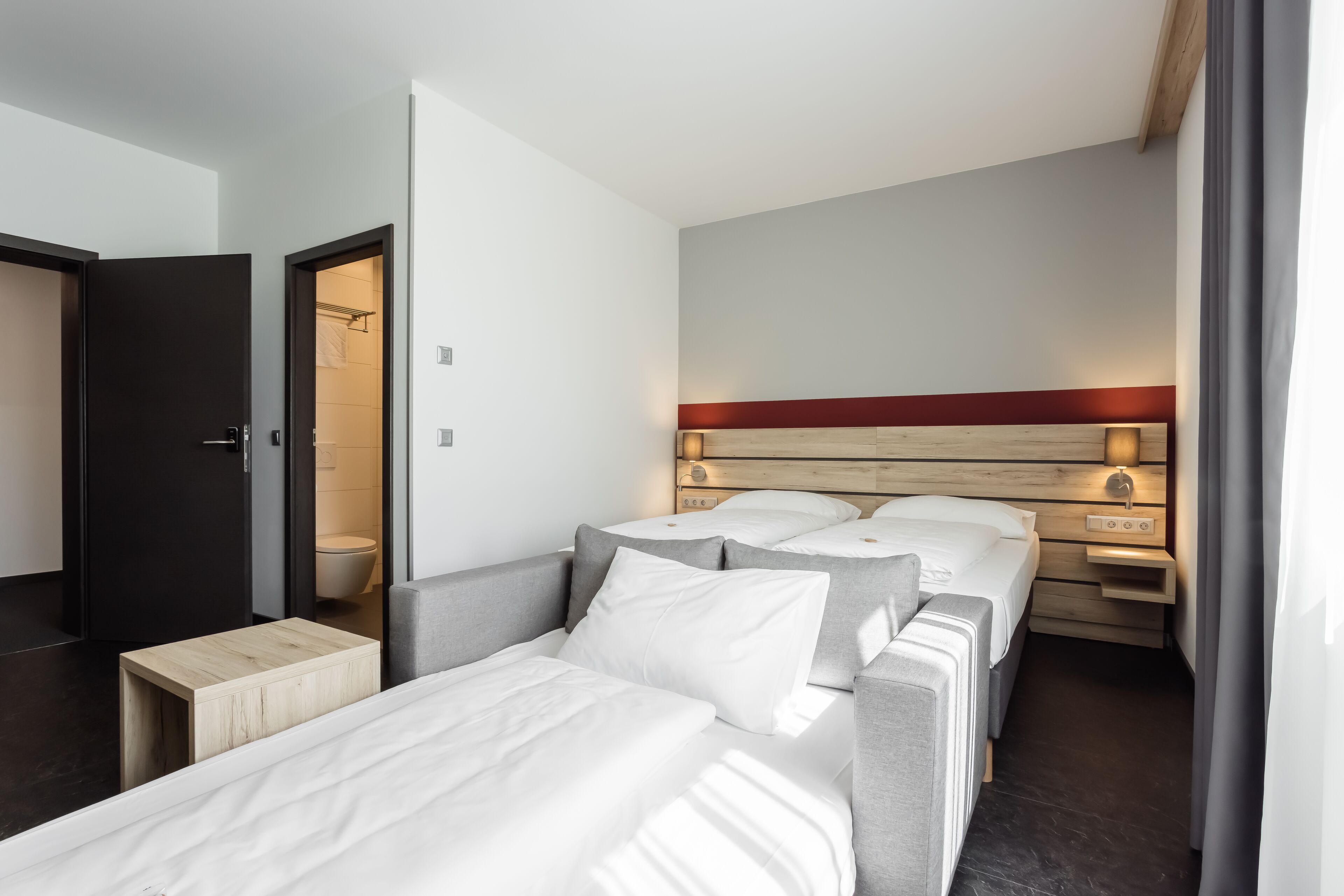 A modern hotel room with a large bed, bedside tables, a wooden headboard, and a small wooden stool.