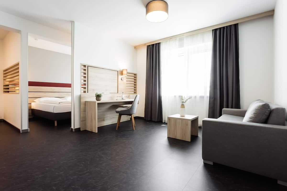A modern hotel room with a bed, a desk, a sofa, and a large window with curtains.
