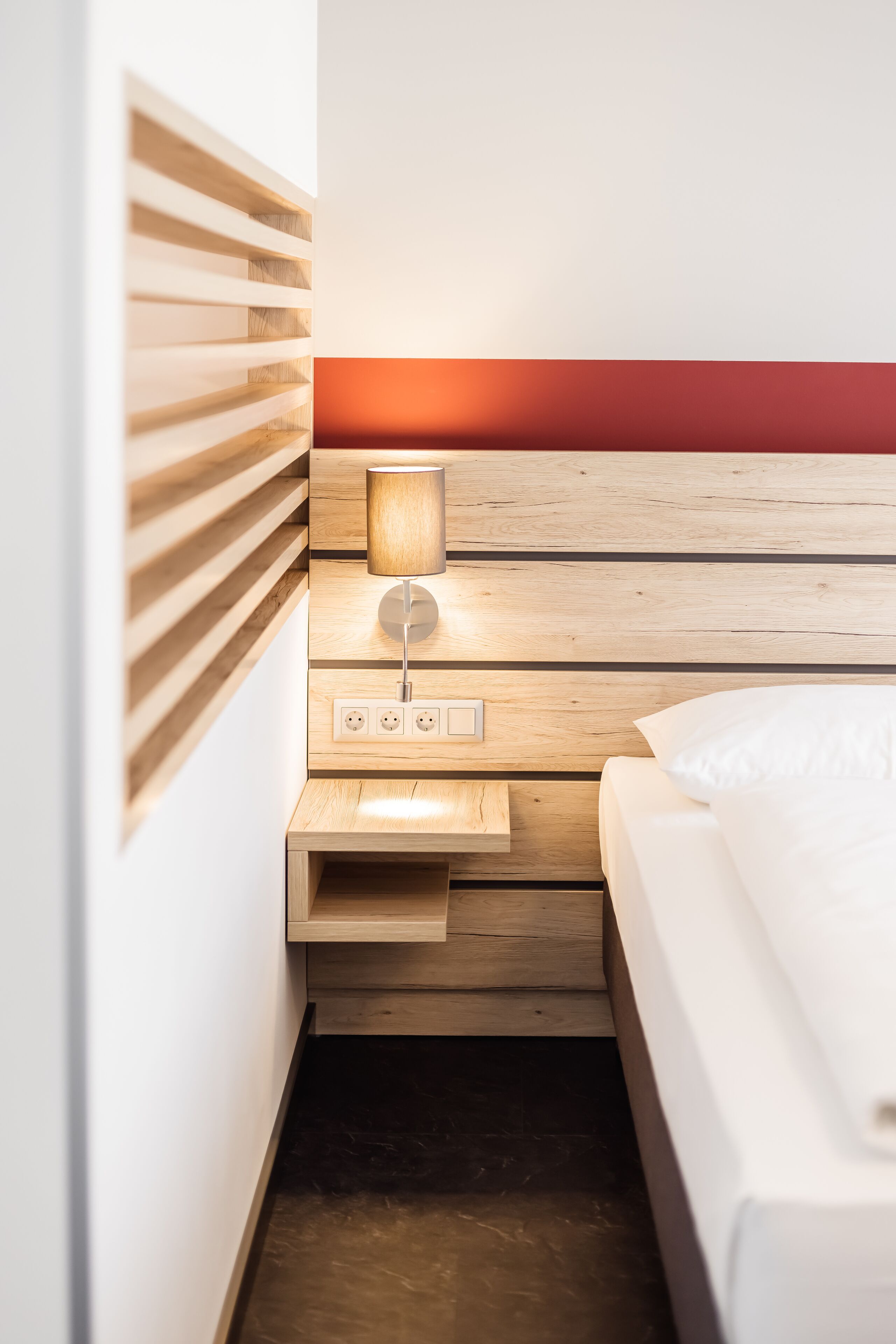 A hotel room with a wooden headboard, a bedside table with a lamp, a power outlet, and a bed.