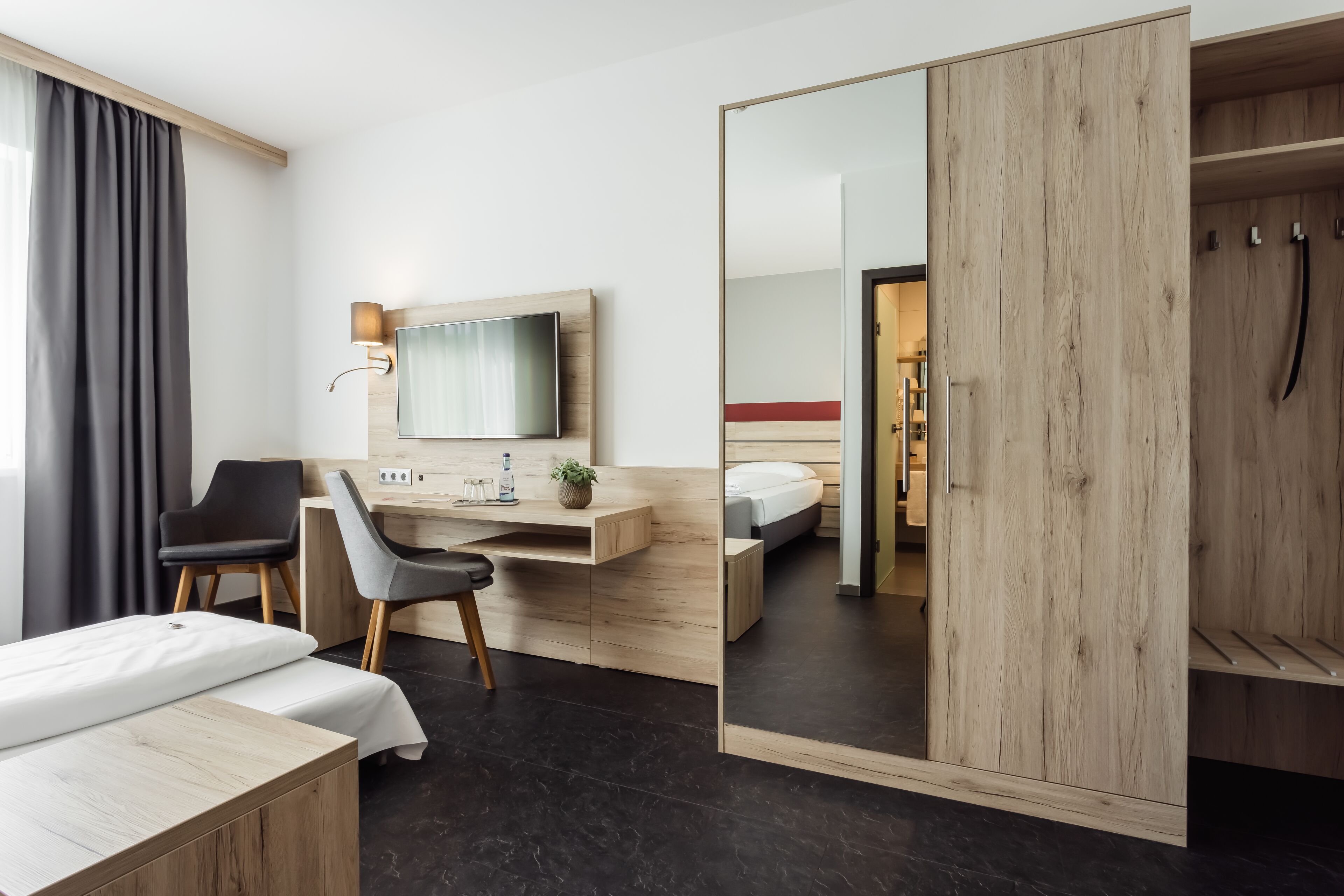 A modern hotel room with a bed, a desk with a flat-screen TV, a chair, a wardrobe, and a large mirror.