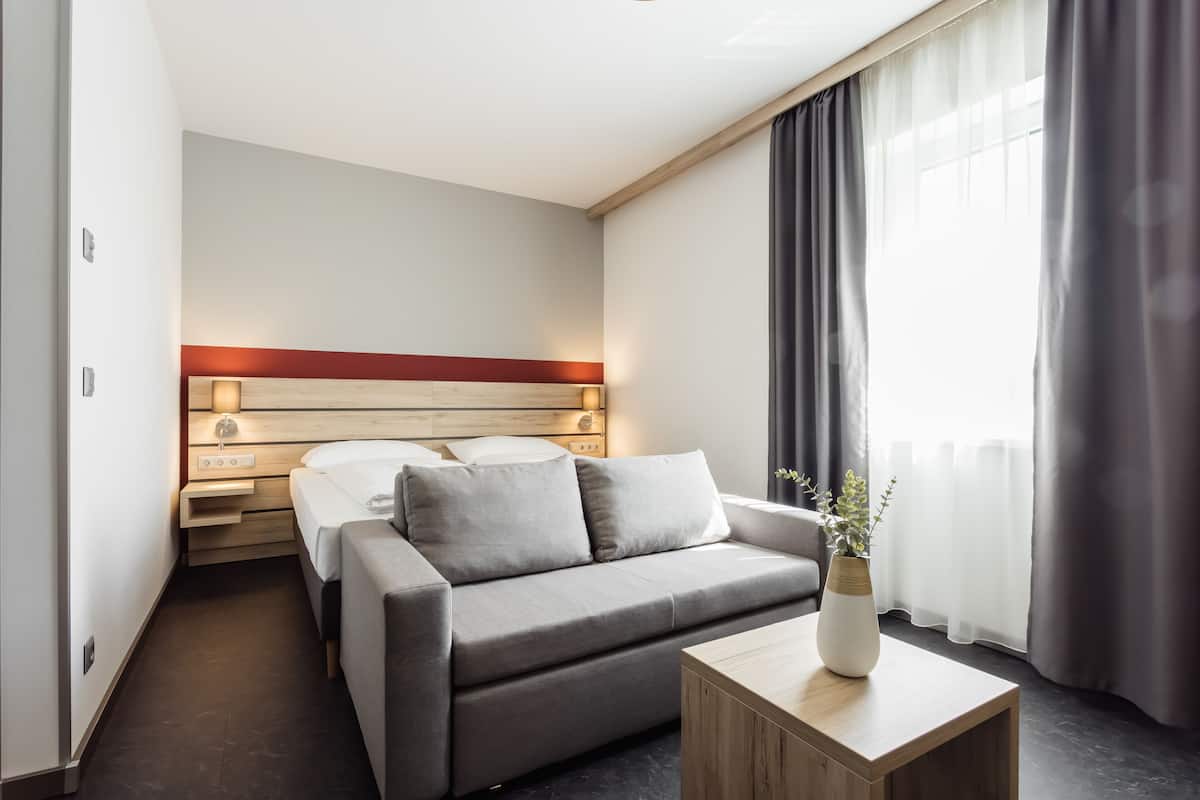 A modern hotel room with a bed, a sofa, a bedside table, and a window with curtains.
