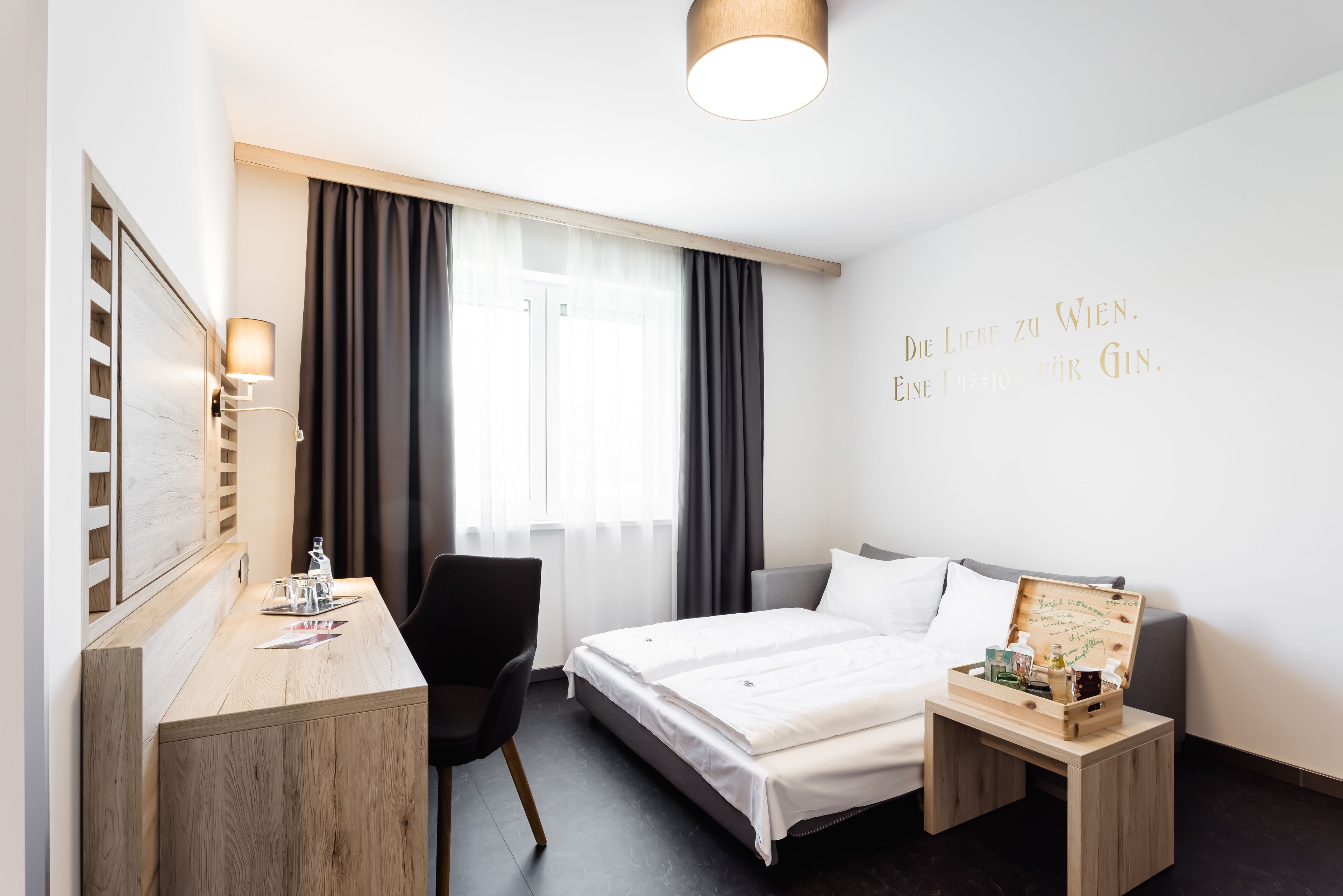 A hotel room with a bed, a desk, a chair, a bedside table, and a wall with a quote.