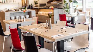 A restaurant with wooden tables and chairs, some with red cushions. There are vases with flowers on the tables.