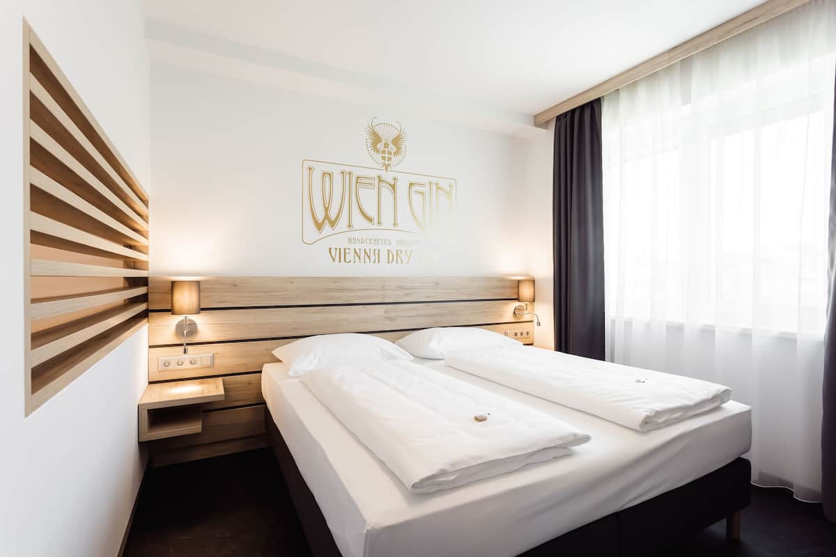A hotel room with a wooden headboard, a bed with white linens, a wall-mounted light, and a window with curtains.