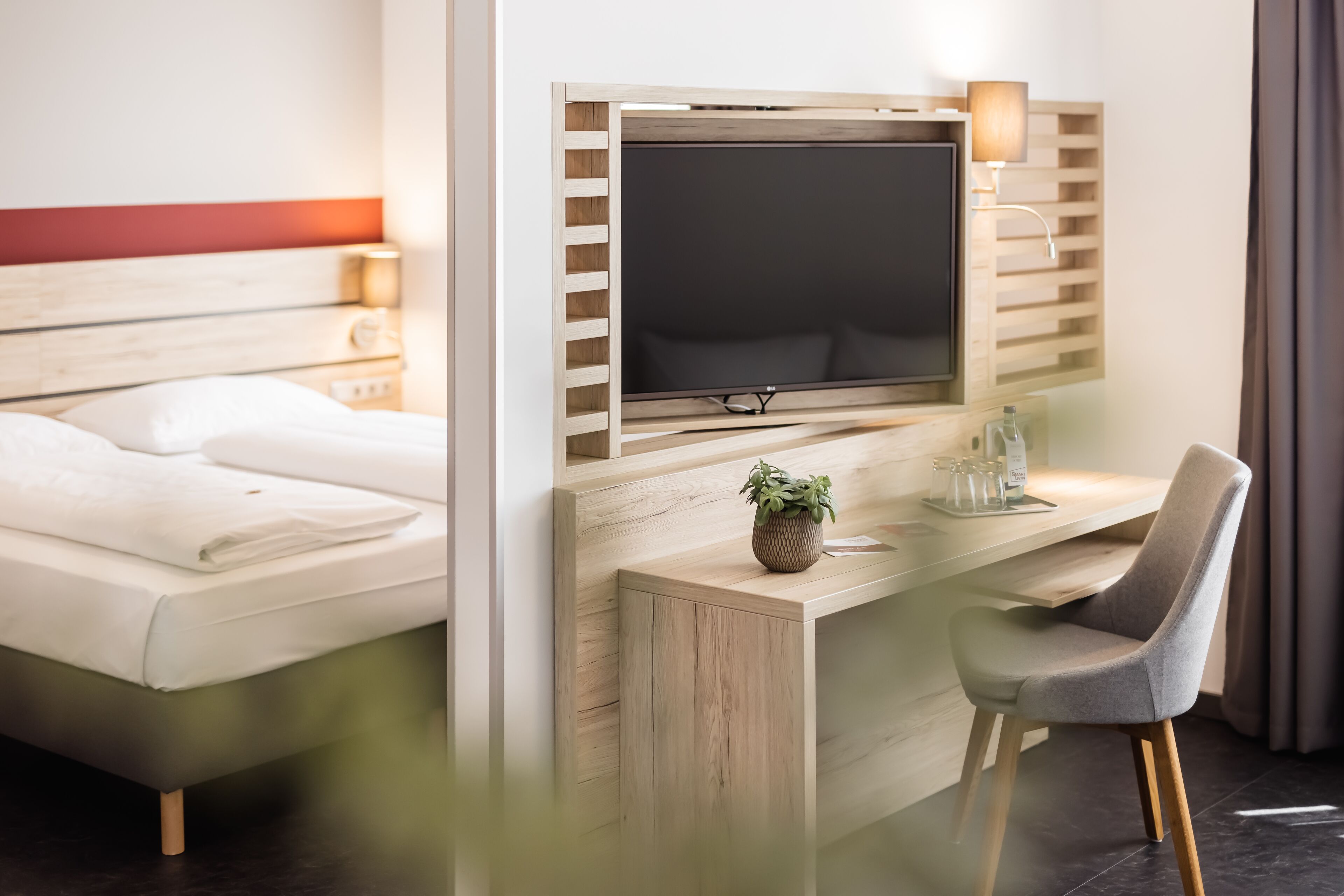 A modern hotel room with a bed, a desk with a flat-screen TV, a chair, and a small potted plant.