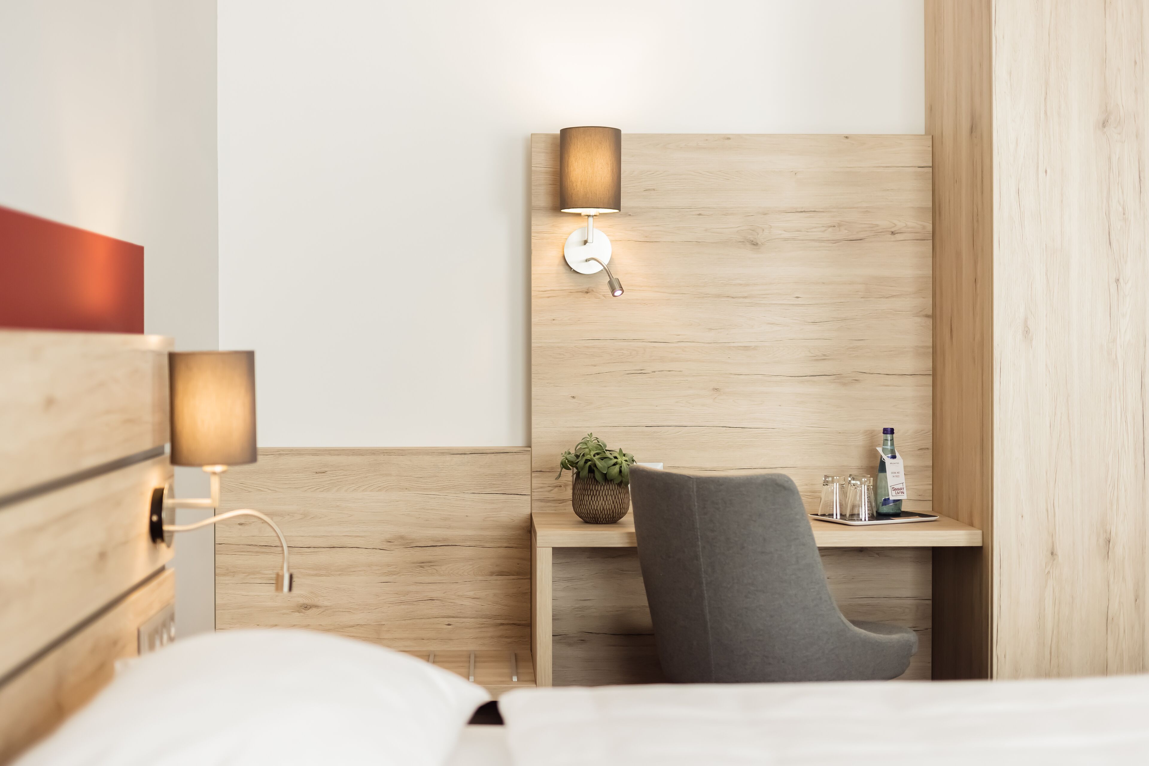 Ein modernes Hotelzimmer mit einem hölzernen Kopfteil, einer Nachttischlampe, einem kleinen Schreibtisch mit Stuhl und einer Pflanze.