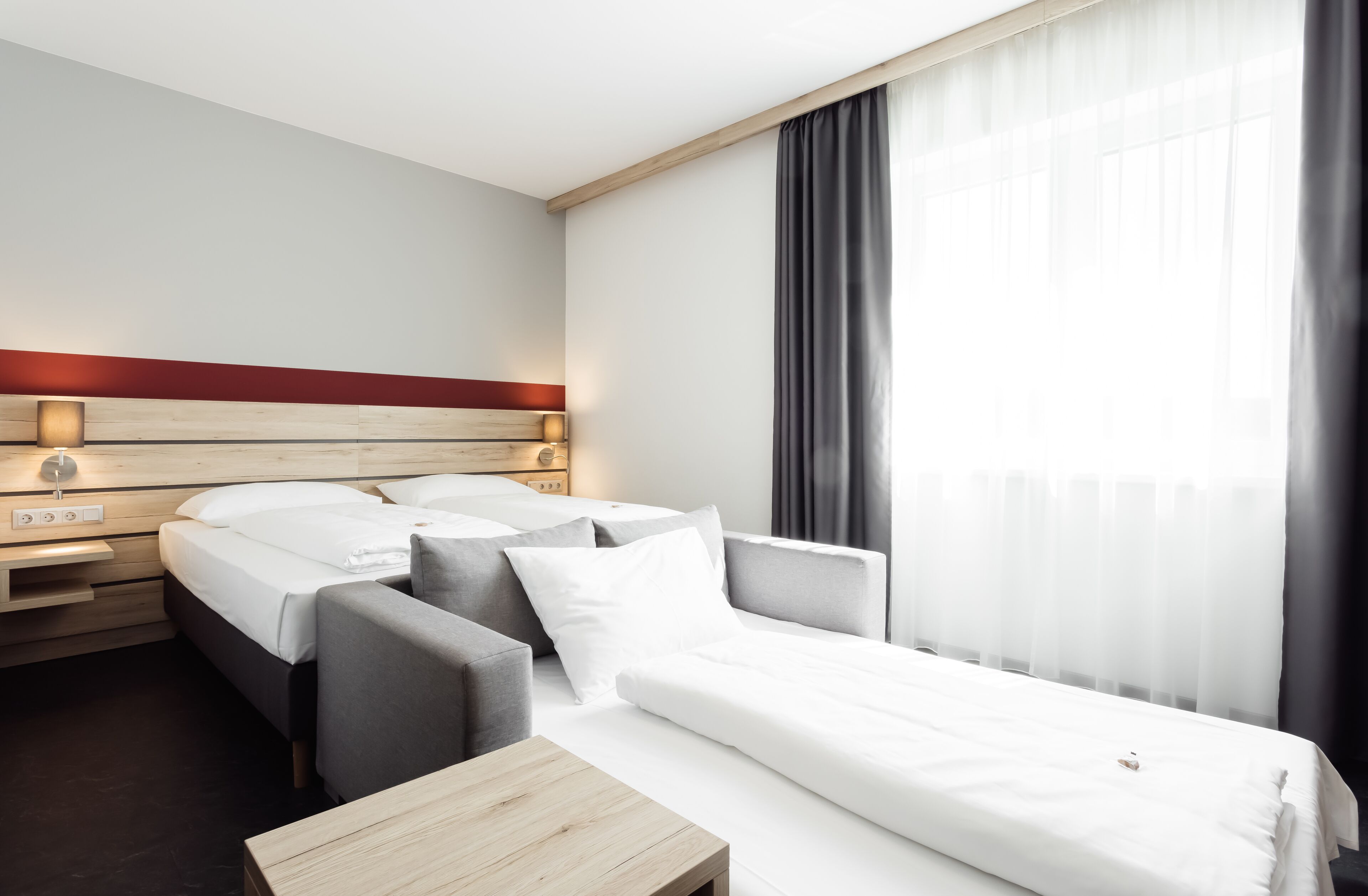 Habitación de hotel con cama, sofá, mesita de noche de madera, lámpara y ventana con cortinas.