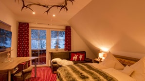 A cozy room with a bed, a desk, and a chair. There is a window with curtains, a fireplace, and a ceiling with antlers. - Hotel Höhengasthof Grüner Baum (Feldberg)