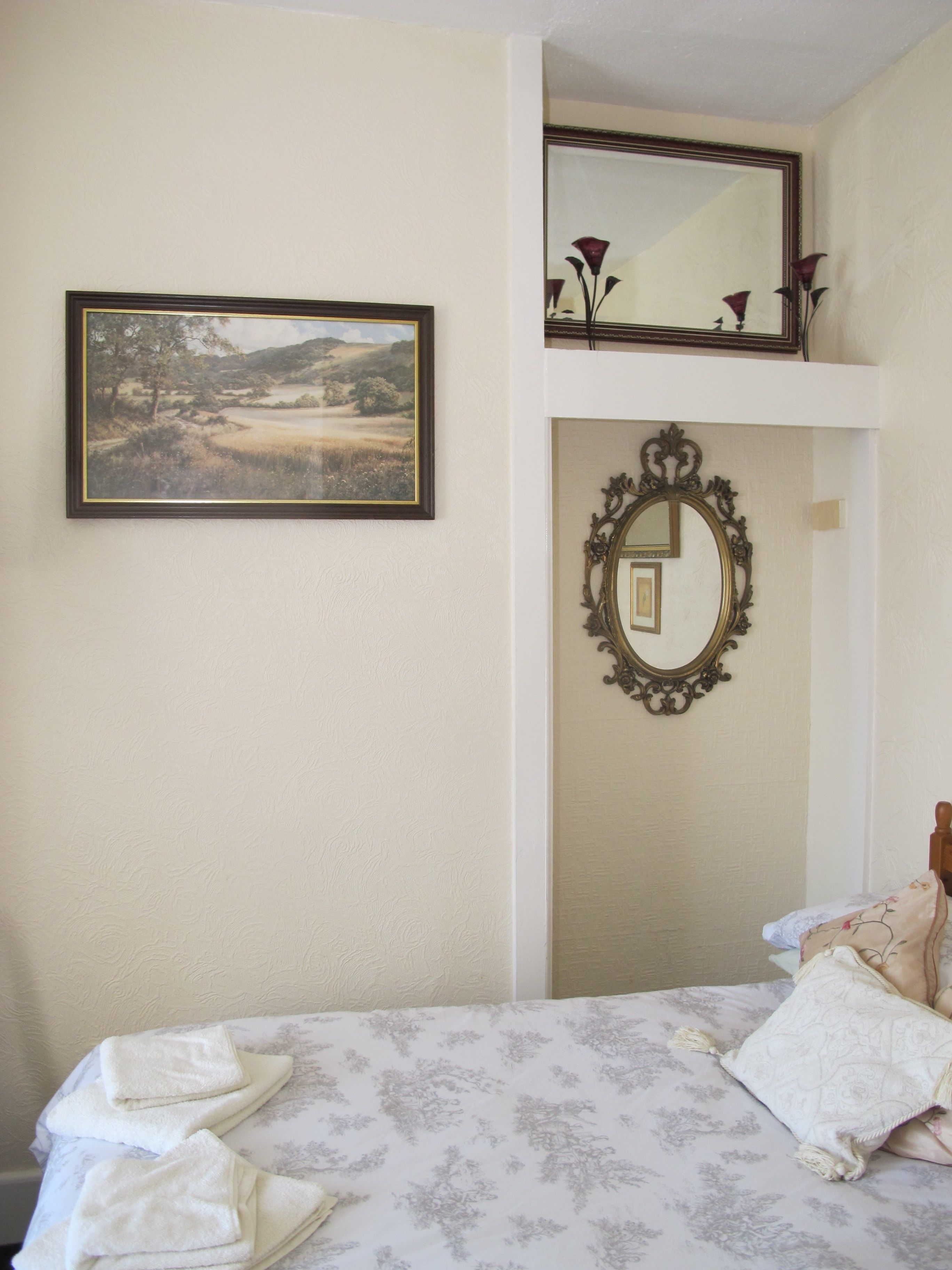Un dormitorio con una cama, un cuadro de un paisaje enmarco y un espejo con un marco ornamentado.