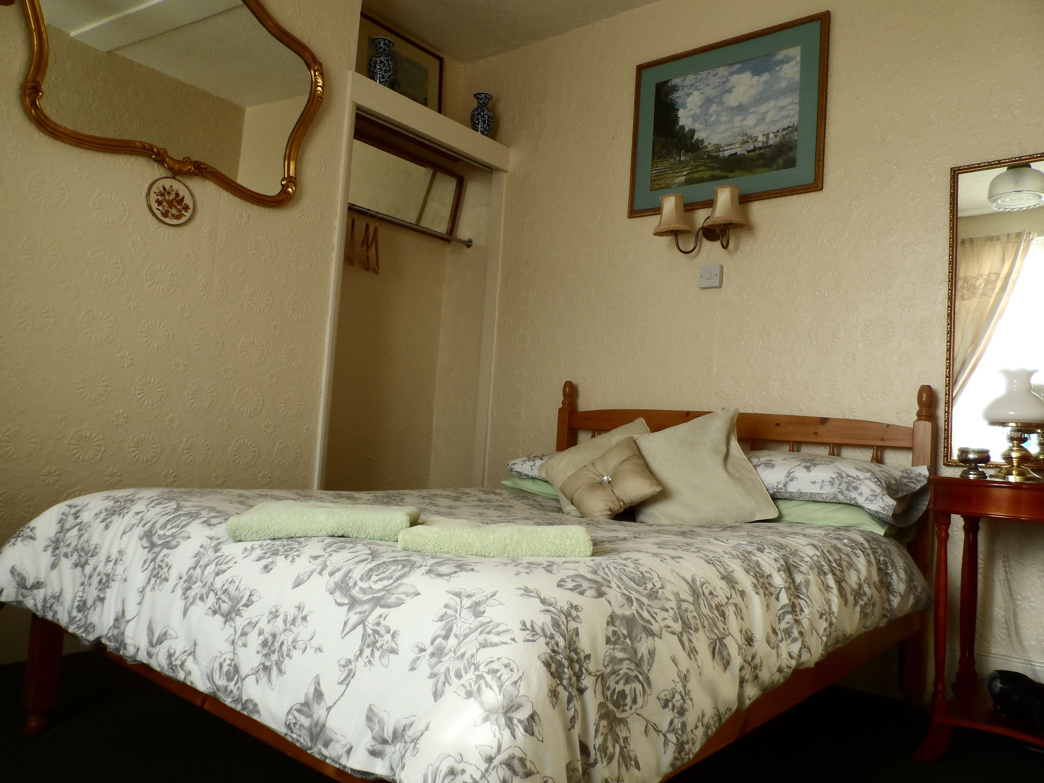 A bedroom with a floral bedspread, a wooden headboard, a bedside table with a lamp, and a framed landscape painting on the wall.