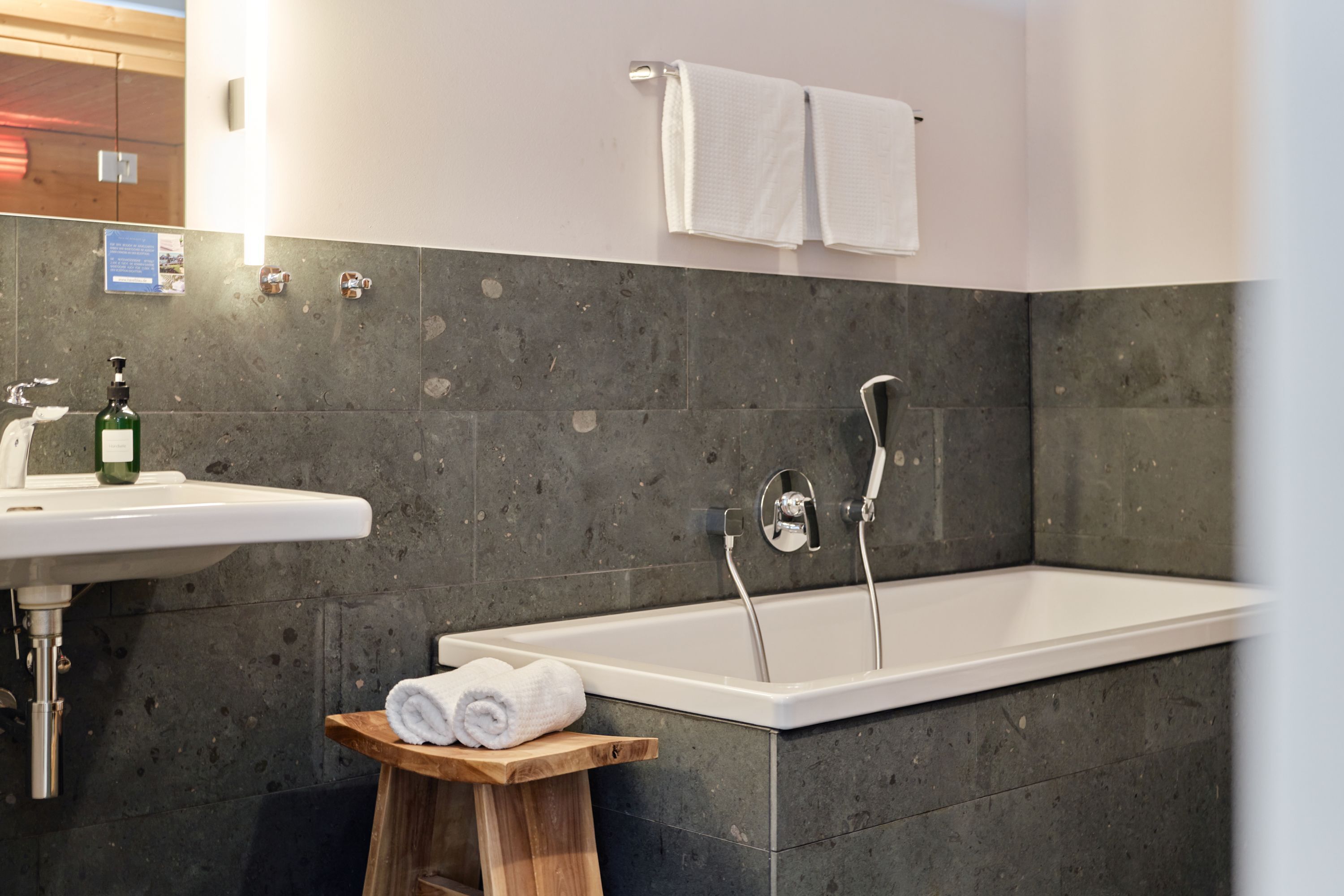 Ein Badezimmer mit einem weißen Waschbecken, einem hölzernen Hocker mit Handtüchern, einer Badewanne und einem Handtuchhalter.