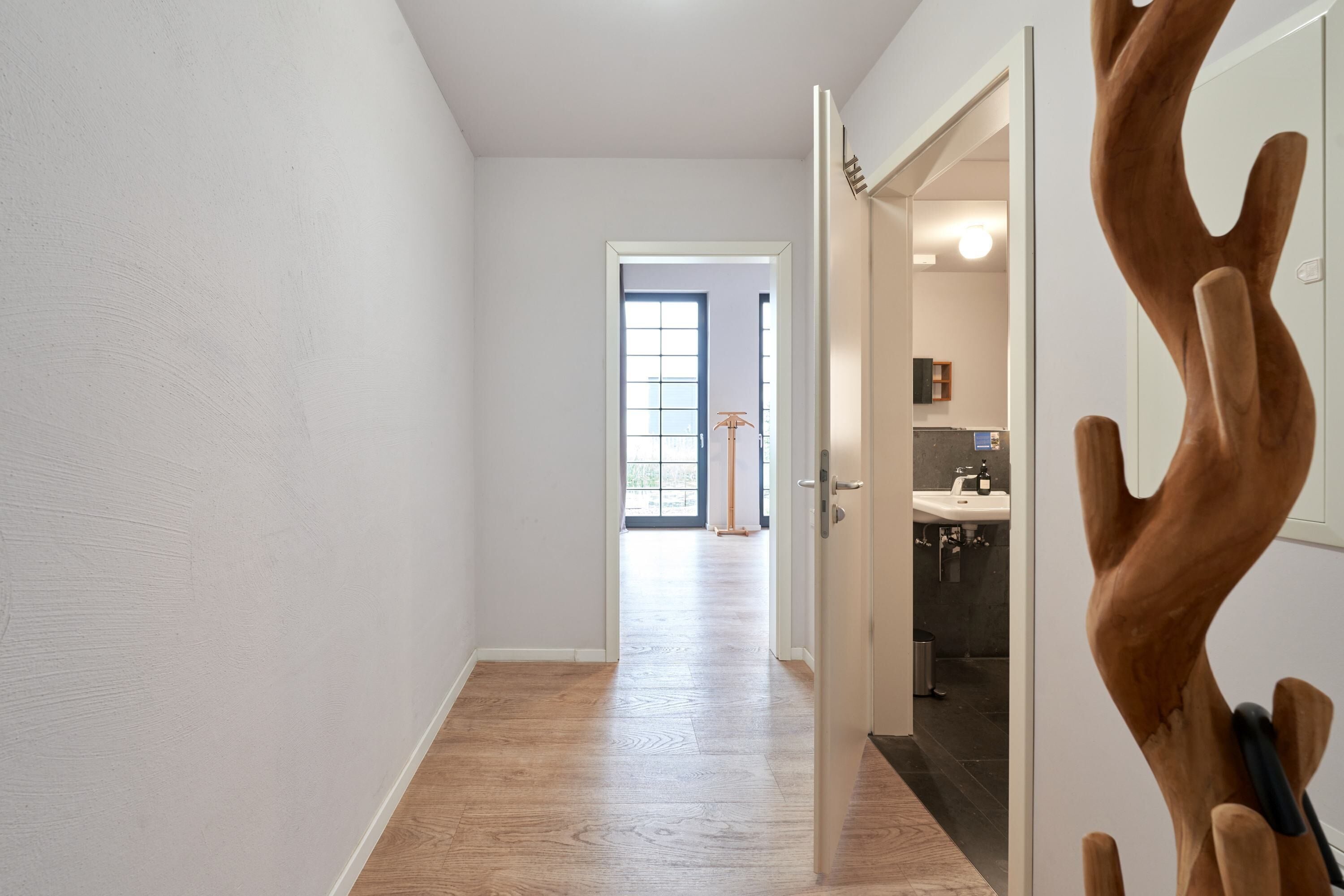 Um corredor com piso de madeira, parede branca e porta que dá para um banheiro com pia e espelho.