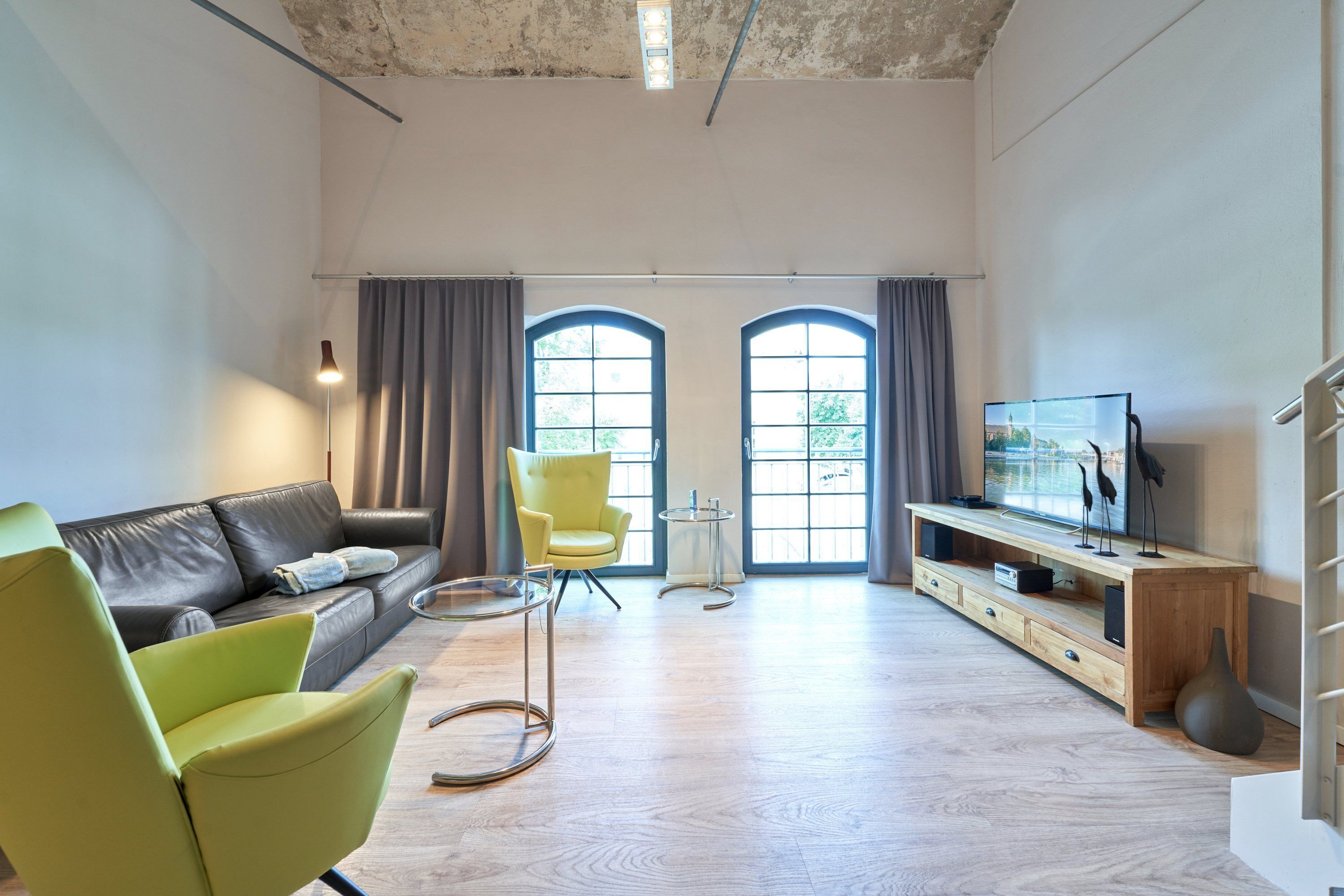 A modern living room with a sofa, a green armchair, a TV on a wooden stand, and a large window with curtains.